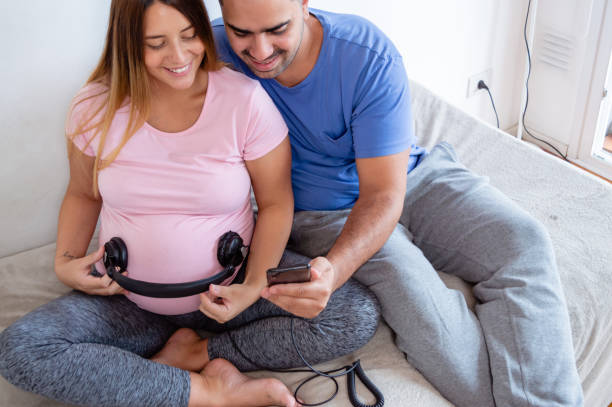 mulher grávida com fones de ouvido em sua barriga e marido tocando música com o telefone para o bebê - heterosexual couple audio - fotografias e filmes do acervo