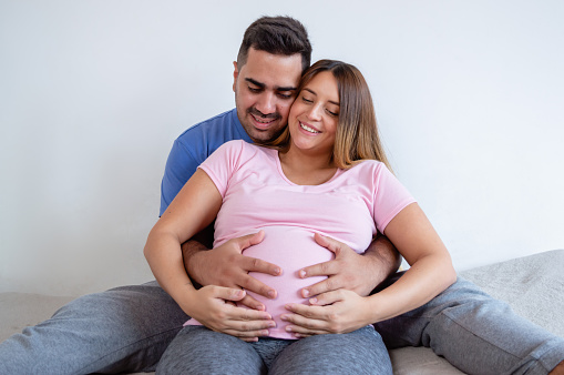 happy caucasian pregnant woman with her husband sitting on bed smiling and touching baby on tummy, doing pregnancy therapy and exercise routine.