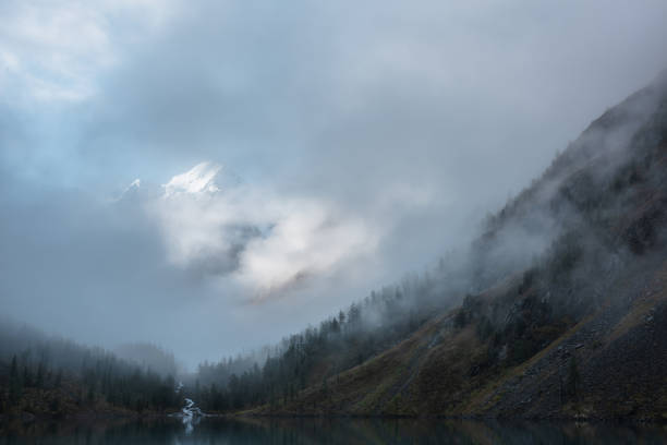 istock Tranquil scenery with snow castle in clouds and alpine lake. Mountain creek flows from forest hills into glacial lake. Snowy mountains in fog clearance. Small river and coniferous trees in low clouds. 1426729146