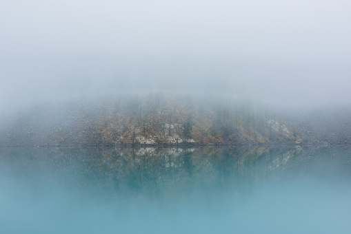 Simple meditative landscape with turquoise lake in thick fog. Tranquil view through clearance in dense fog to steep slope in fading autumn colors and mirror mountain lake. Reflective azure alpine lake