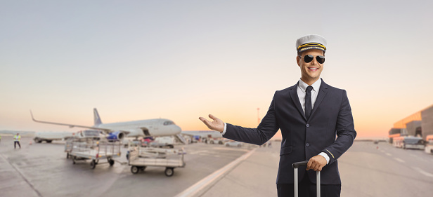 Pilot standing with a suitcase on an airport with plane in the background and pointing with hand