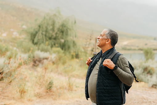 Nature, health and a man hiking in the countryside, walking with backpack and smile. Happy, calm, and on retirement, a senior on a travel adventure, trekking in a forest landscape with trees and lake