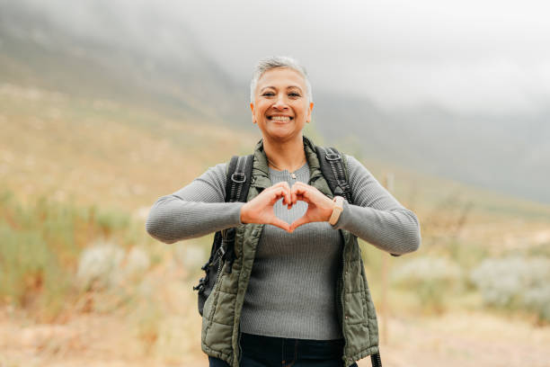 Nature hiking senior woman with love and heart sign or hand for healthy, wellness and care on adventure walk on hill, green mountain environment. Trekking person with emoji hands, portrait and smile stock photo