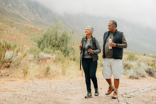 Nature, health and a couple hiking an adventure trail during retirement. Happy, a man and woman walking in a natural landscape and drinking water. Seniors on a weekend walk on a cloudy mountain path.