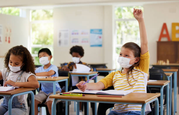 istock Wearing face mask to protect from covid while learning in class, answering education question and studying with students in a classroom. Girl sitting at a desk and raising hand during a pandemic 1426755574