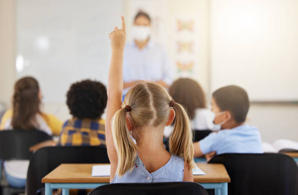 Learning, smart and little girl in class holding up a hand to answer a question at school. Back view of a young student sitting at a classroom desk looking to solve the questions a teacher is asking aprendiendo, inteligente y niña en clase levantando una mano para responder una pregunta en la escuela. vista trasera de un joven estudiante sentado en el escritorio de un aula buscando resolver las preguntas que un maestro está haciendo