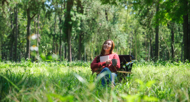 Portrait of a beautiful young asian woman reading a book and drinking coffee while sitting on a camping chair in the park stock photo