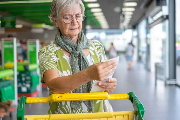 Photo of Senior woman in the supermarket checks her grocery receipt looking worried about rising costs - consumerism concept, rising prices, inflation