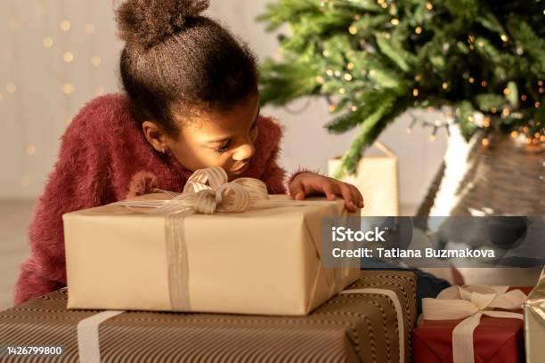 Nine-year-old African-American girl sitting at home under the Christmas tree with gifts.The girl is wearing a pink sweater and jeans.New Year and Christmas concept.Diverse people.