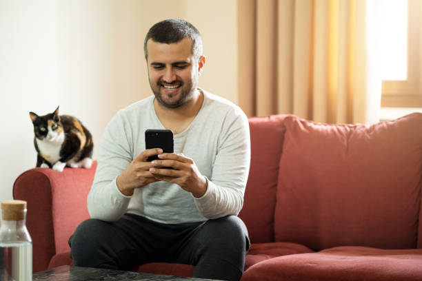 handsome short haired man holding smartphone and sitting on the couch with his cat. copy space - sexting imagens e fotografias de stock