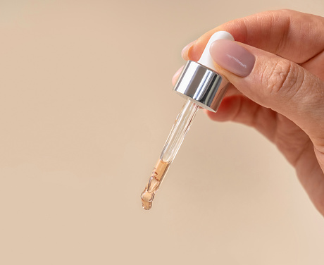 Female Hand holding pipette with drop falling down over light beige close up. Natural skincare product, serum or essential oil. Cosmetic routine.  Medical research