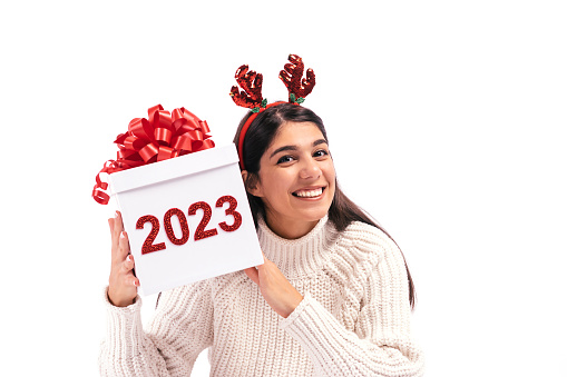 Young dark-haired woman on a white background holds a gift box with the numbers 2022 in her hands.Woman is wearing a Santa Claus hat,white t-shirt and shirt.Concept of the New year 2023.Copy space.