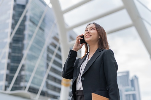 Portrait moment photo of an attractive young asian professional businesswoman lady with nice smile on her face talking on the phone while walking and looking up the sky and holding a clip folder board in a central business district in Bangkok city