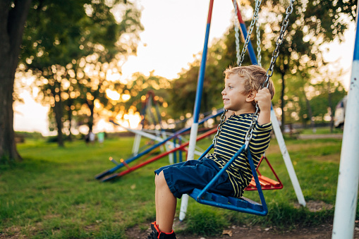 Side view of an adorable little boy is swinging on a swing at a children's playground