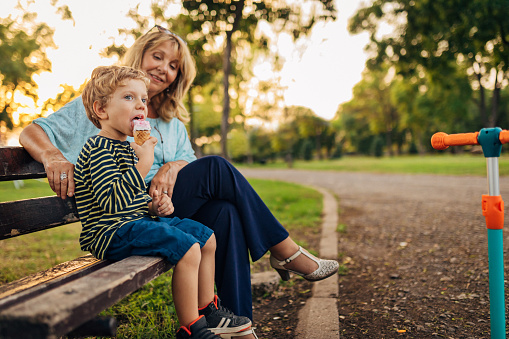 Cheerful boy eating his favorite ice cream while sitting on a park bench with his grandmother