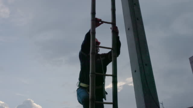 A telecoms worker is shown working from a utility pole ladder while wearing high visibility personal safety clothing, PPE, and a hard hat.