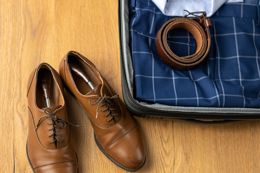 Male leather shoes and formalwear in suitcase close up, business travel preparation. Luggage packing, businessman clothing on wooden floor