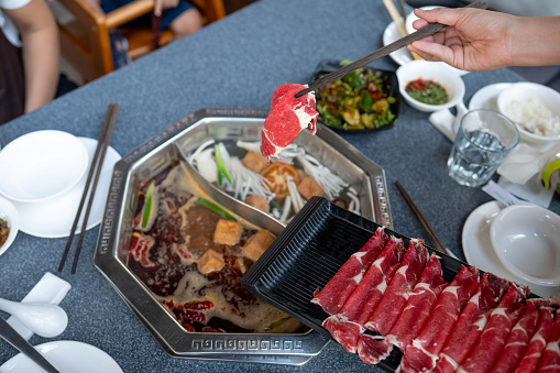 Person adding raw beef in a boiling hot pot with lots of ingredients to be cooked in a boiling soup in hot pot style Chinese restaurant