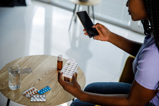 Copy space shot of young Black woman sitting at coffee table in her living room, holding a pill organizing case and reading her medical prescription via smart phone.