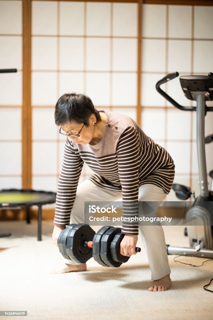 Senior Japanese Woman Exercising At Home Doing Squats With A Dumbbell