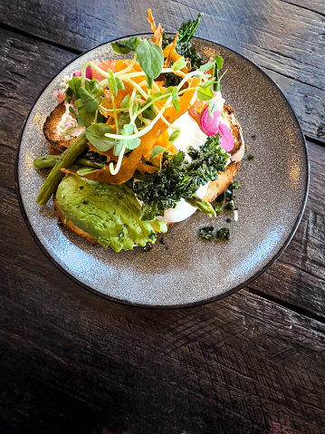 Vertical high angle closeup photo of poached free range eggs on slices of toasted sourdough bread with smoked salmon, sliced avocado, asparagus, pickled onion, baked sweet potato and kale crisps and a micro herbs garnish on a round ceramic plate on a rustic wooden table in a cafe.