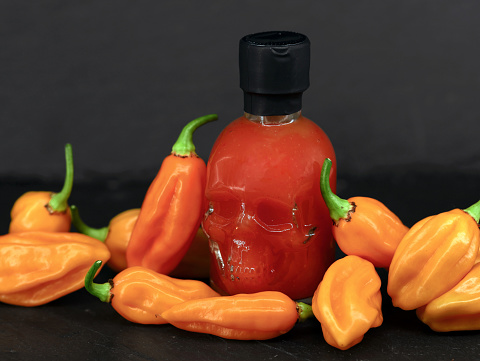 Chili sauce bottle in the shape of a skull among hot orange chili peppers on a black background.