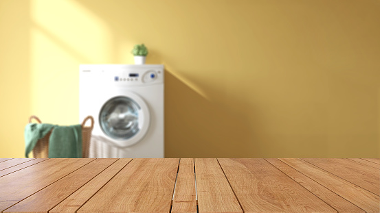 Wooden tabletop or countertop in laundry utility room with white washing machine, basket and vibrant clean yellow wall with sunlight at home for household washing and cleaning product display
