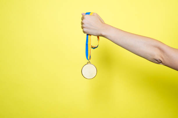 黄色の背景に隔離された黄色の青いリボンに金メダルを手持ちします。メダルモックアップ - gold medal medal ribbon trophy ストックフォトと画像