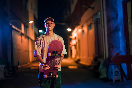 portrait asian Chinese young men standing at back alley holding skateboard looking at camera smiling