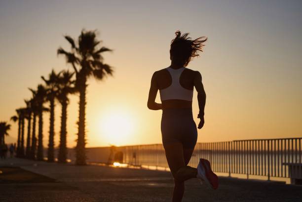 Woman running jogging at sunrise sunset silhouetted with palm trees and ocean in background stock photo