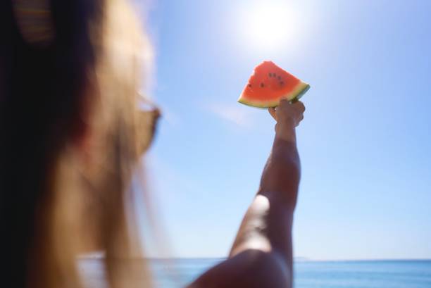 Woman's hand holding slice of watermelon on the beach on a sea background. Tropical fruits, healthy food, summer. Sweet, juicy treats in the dish. Healthy eating and lifestyle. Food concept stock photo