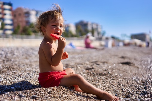Adorable Young Toddler Eats Delicious Ripe Watermelon by the Ocean on Beautiful Beach During the Summer