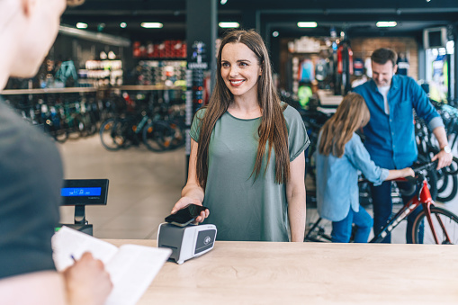 young woman paying at checkout with mobile phone in bike shop, husband and daughter  with new bicycle in background
