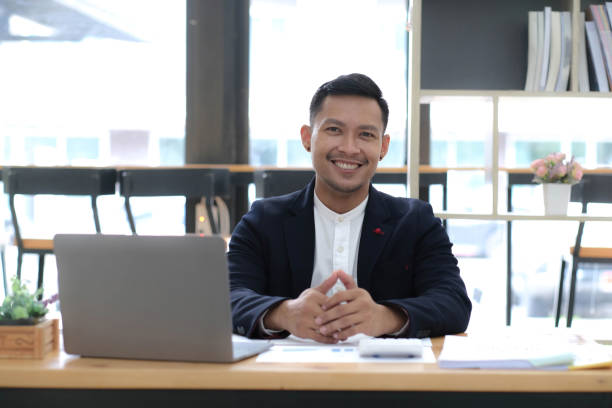 istock Portrait of a young Asian businessman smiling while using a laptop and writing down notes while sitting at his desk in a modern office 1426944891