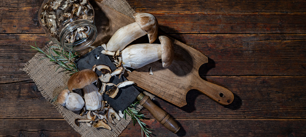 Dark food photography background - Fresh and dried forest mushrooms / Boletus edulis (king bolete) / penny bun / cep / porcini / mushroom, knife and rosemary herbs on cutting board on table, top view