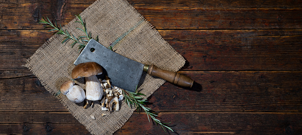 Dark food photography background - Forest mushrooms / Boletus edulis (king bolete) / penny bun / cep / porcini / mushroom, knife and rosemary herbs on jute sack on table, top view