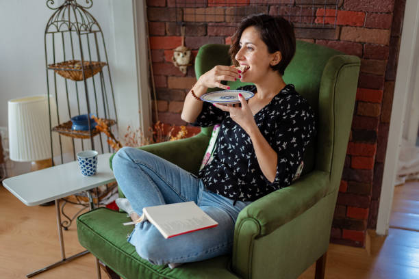 Woman sitting on couch in living room eating vegan snack Woman sitting on couch in living room eating vegan snack Smart Snacking a Habit stock pictures, royalty-free photos & images