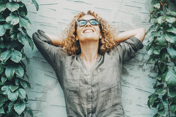 mujer feliz y satisfecha sonriendo y mirando al aire con hiedra verde de la naturaleza a ambos lados. retrato de mujeres alegres de pie frente a una pared gris al aire libre. concepto de felicidad. una persona - menopausia fotografías e imágenes de stock