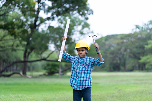 African American child boy in engineering holding construction blueprint and steel hammer wearing yellow safety helmet outdoor in the park. Education concept