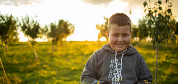 The boy looks at the camera and smiles. The boy poses in a village orchard, behind him is a beautiful sunset. Golden hour
