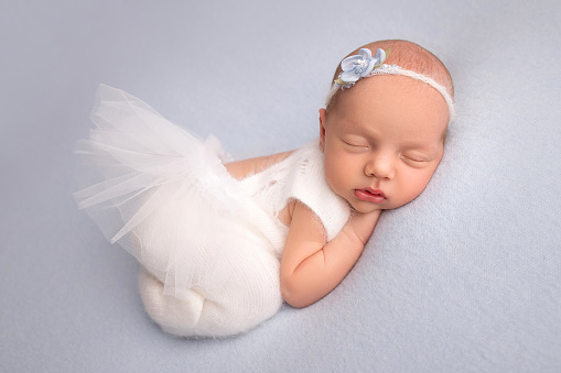 Sleeping newborn girl in the first days of life in a white ballet dress with a white bandage and a blue flower on a blue background. Studio macro photography, portrait of a newborn ballerina