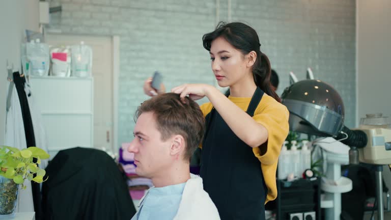 Young female hairdresser using hair dryer and round brush while drying customers hair