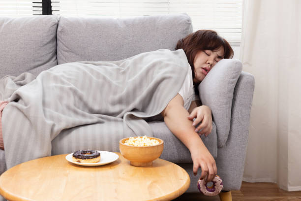 leniwa kobieta z nadwagą śpi na kanapie z pączkiem w ręku - jałowy zdjęcia i obrazy z banku zdjęć