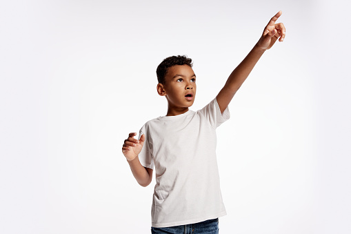 Shocked african little boy in white tee pointing and looking up isolated over white background. Kids emotions, studying, child psychology and ad concept