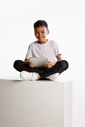 Remote studying. Curly school age boy in casual tee and jeans sitting on big box with digital tablet isolated on white background. Happy, joyful, childhood, kids fashion, emotions, facial expression