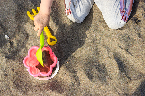 Little kid plays with sand on the beach. Close up little girl hand with toy shovel and bucket. Little girl is filling sand into bucket.
