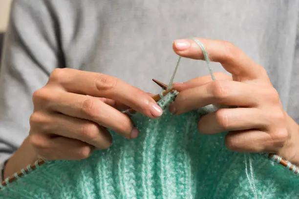 Close up of female hands holding bamboo knitting needles and knitting green woolen sweater. Hobby, relaxation, mental health, sustainable lifestyle.