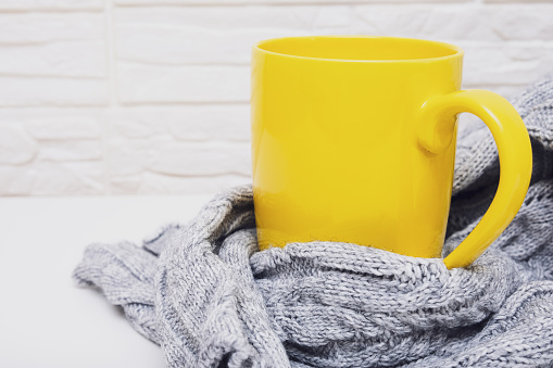 Autumn mood. Yellow cup of hot tea or coffee with a cosy knitted sweater against white backdrop with copy space.