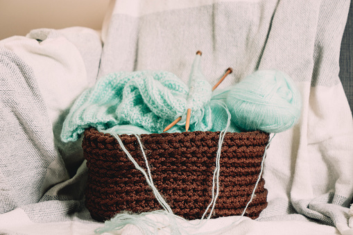 Knitting background. Green Knitting yarn, bamboo needles and new started knitted sweater in crocheted basket on sofa at home. Handmade hobby, relaxation at home, mental health, sustainable lifestyle.