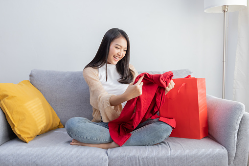 Happy Asian Woman Looking at Her New Clothes From the Shopping Bag  At Home On Couch